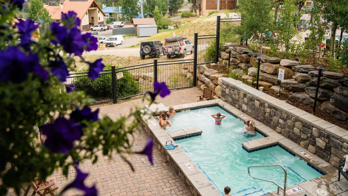 This exterior shot captures a sparkling swimming pool bordered by stone walls and flowering plants. Guests are seen lounging, swimming, and socializing under the warm sun, evoking a sense of leisure and vacation atmosphere. The pool area is inviting and ideal for both relaxation and family fun.