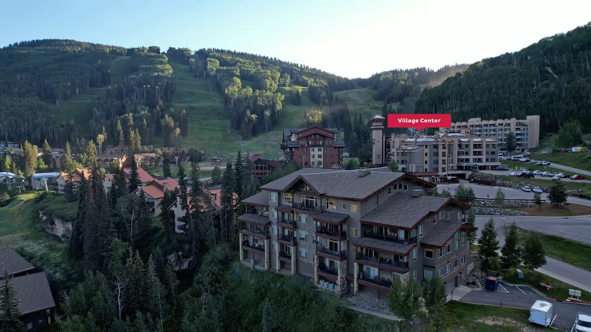 An exterior view of the Village Center buildings beneath a dramatic mountain backdrop, with the Village Center sign prominently displayed. The scene conveys a sense of arrival and the proximity of the resort to nature and village amenities.