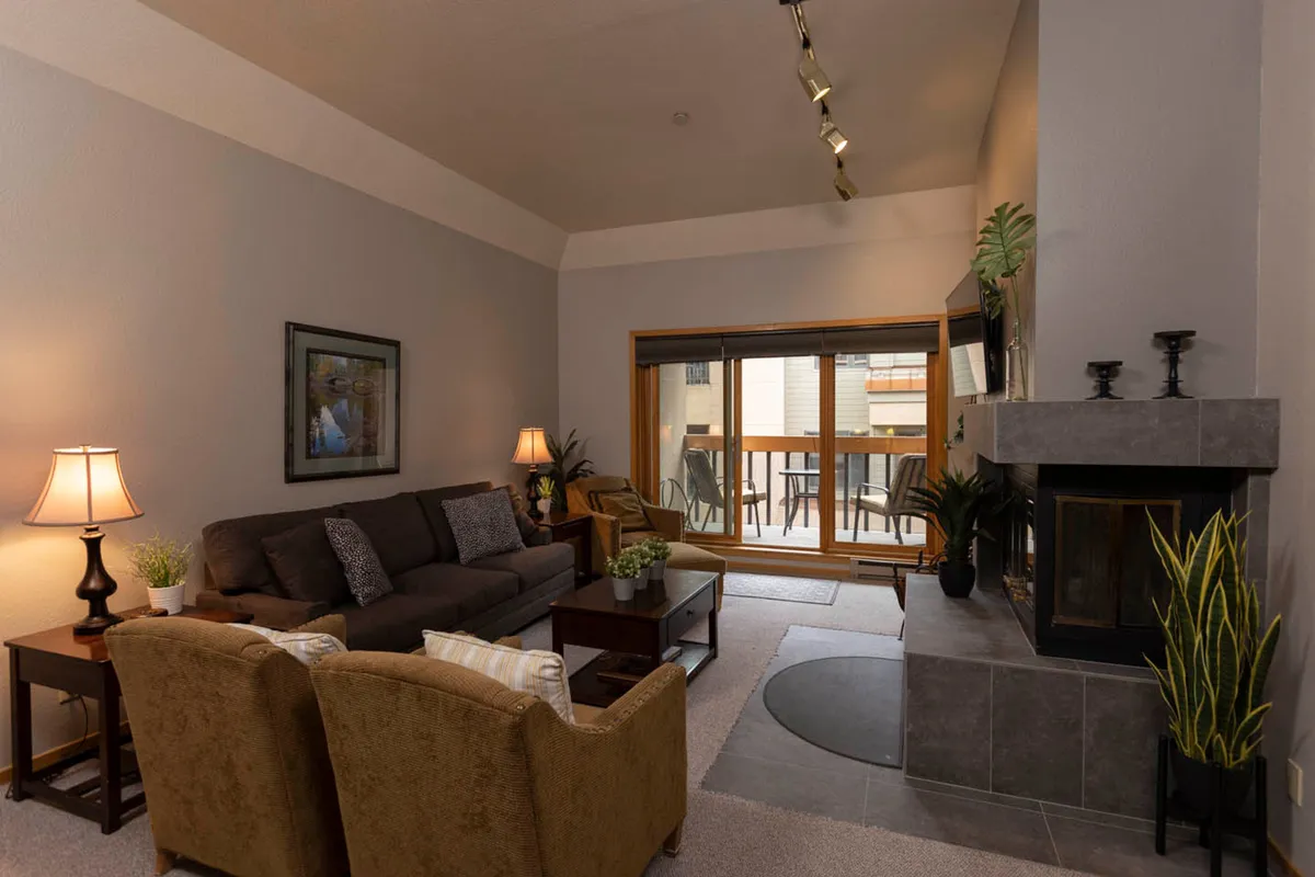 A welcoming living area featuring a comfortable sofa arranged around a stone fireplace, with natural light streaming in from a large sliding glass door that hints at access to the outdoors. The space feels warm and inviting, perfect for relaxing after a day of mountain adventures or gathering with friends and family.