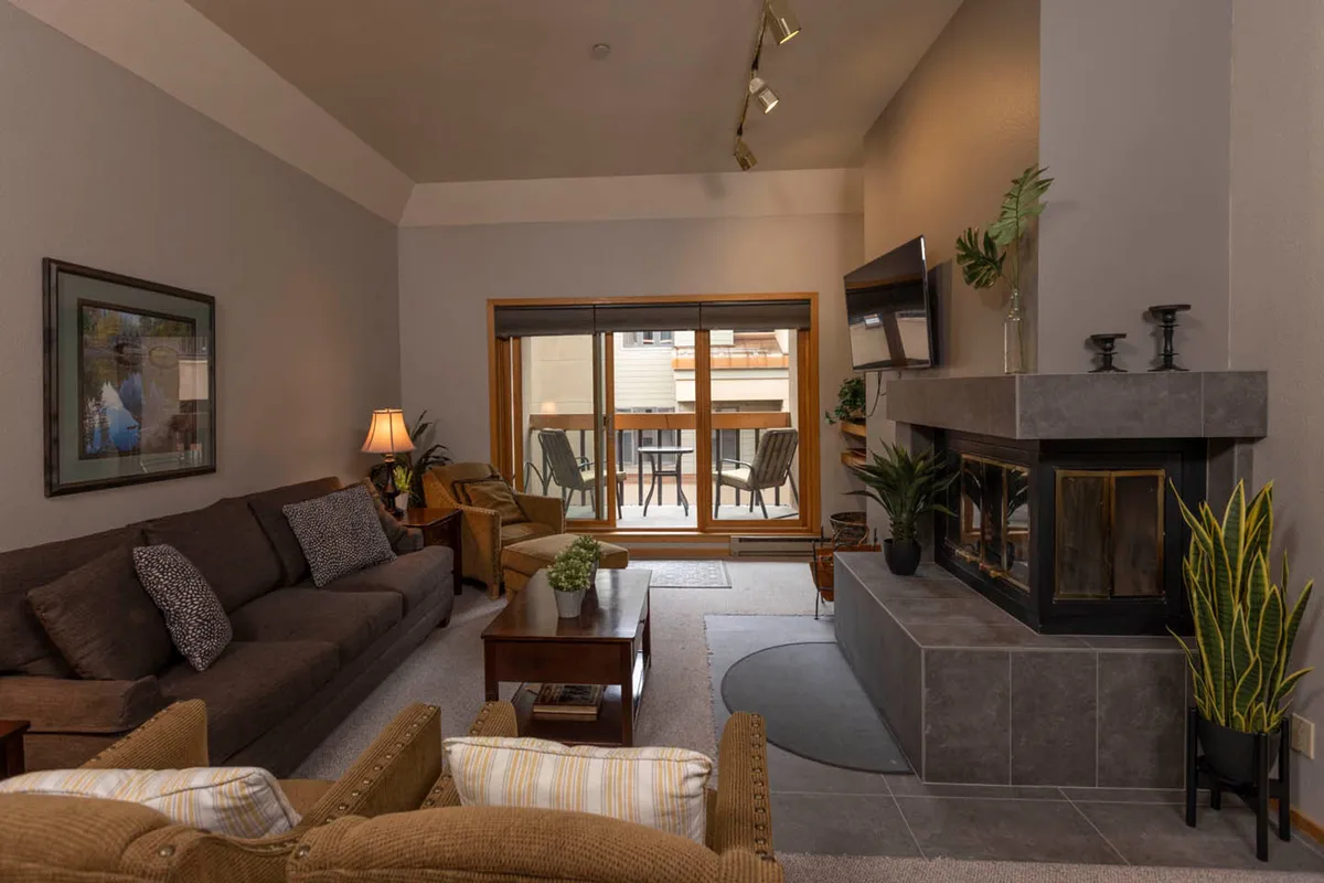 The image features a spacious living area with a prominent fireplace, large windows framing sweeping mountain views, and a comfortable seating arrangement that invites relaxation and socializing. The abundance of natural light highlights the cozy atmosphere and showcases the heart of the gathering space.