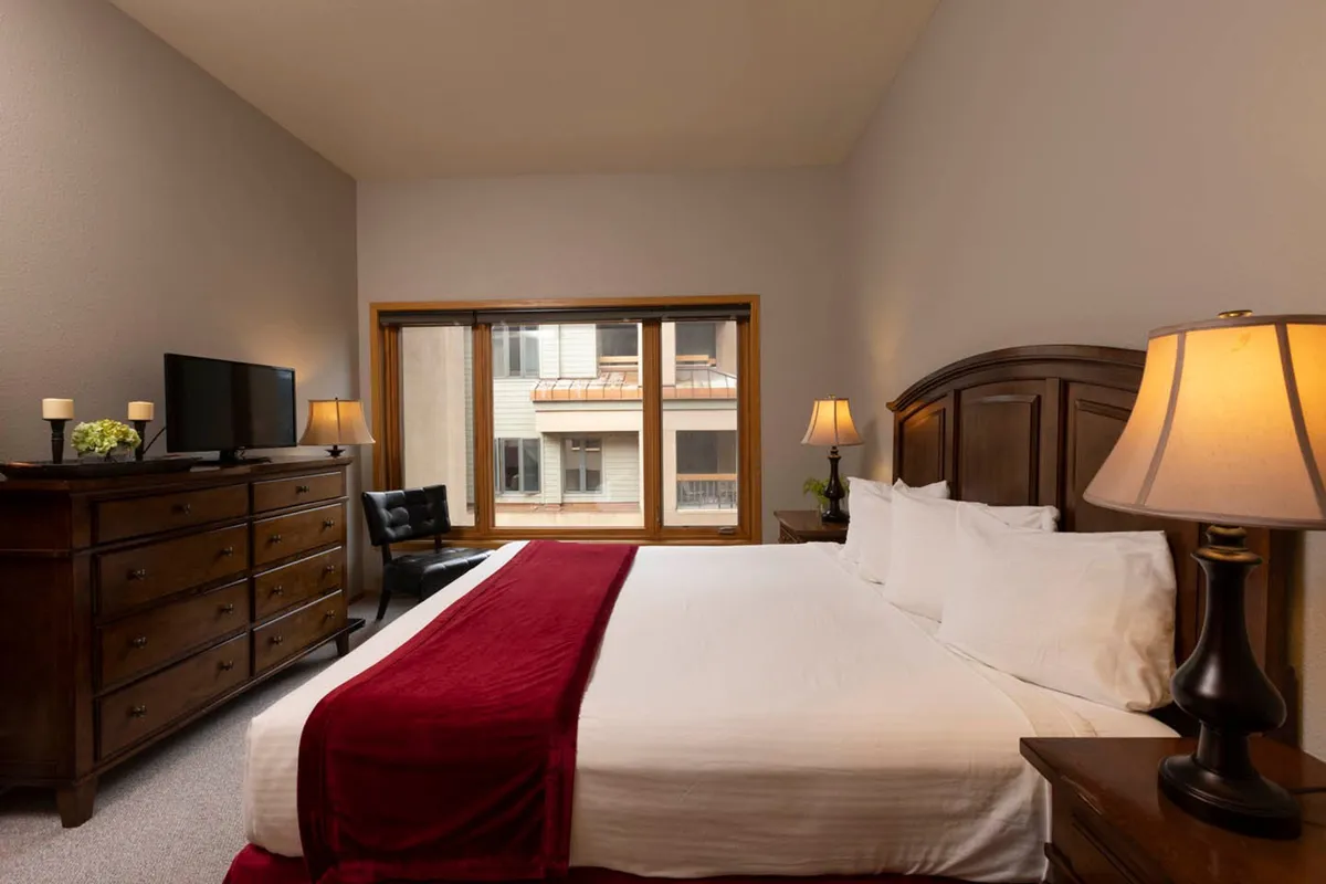 A cozy bedroom featuring a neatly made bed, soft bedding, a window letting in natural light, a dresser, and bedside lamps. The space is calm and welcoming, ideal for relaxation or unwinding with a book or TV after a day outdoors.