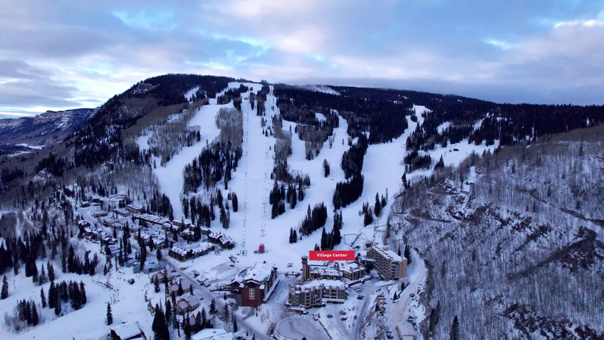 A vibrant winter scene featuring the snow-covered ski slopes, the bustling Village Center, pine trees, and residential resort buildings nestled at the base. The image captures both the adventure of daytime activities on the slopes and the coziness of evening gatherings with the sunrise illuminating the mountains.