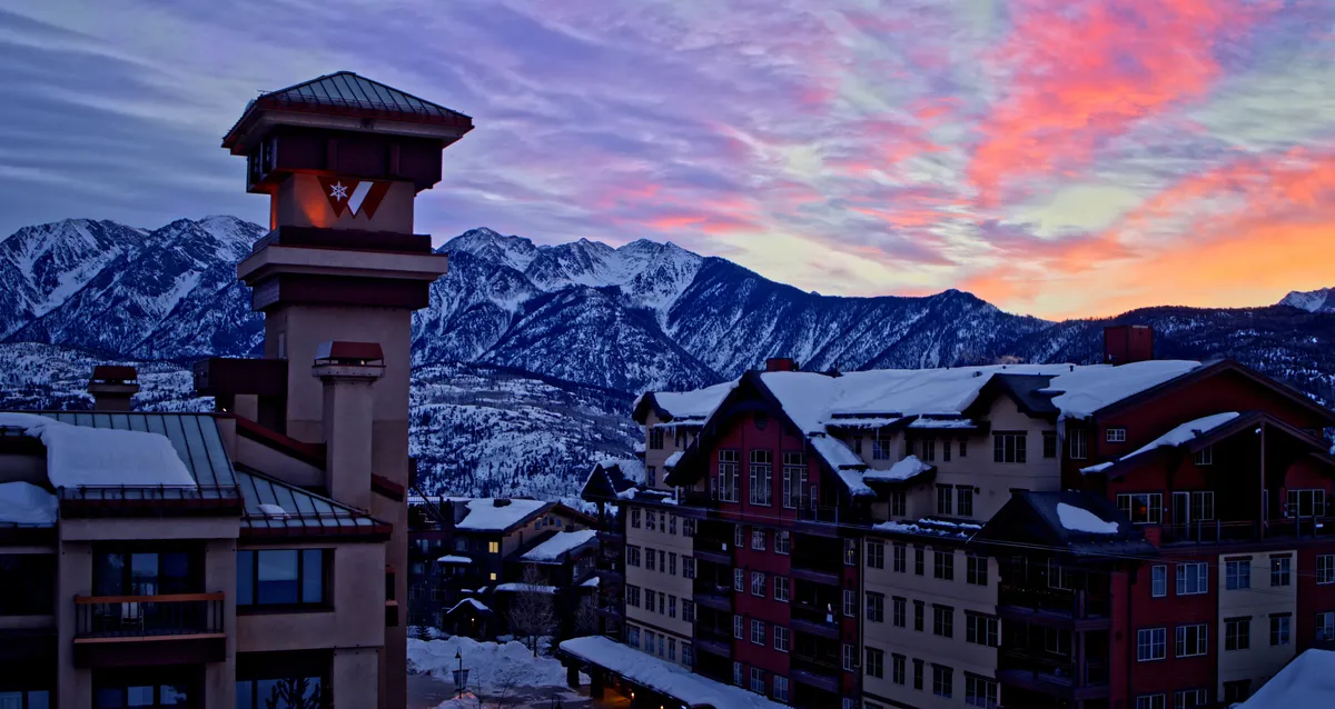 A serene exterior scene showing snow-covered buildings against a backdrop of majestic mountains and a vibrant sunset sky. The cozy chimney hints at warmth inside, while the snow invites thoughts of winter sports and family fun.