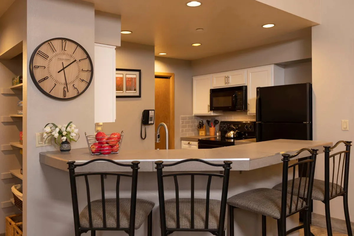 A spacious interior scene featuring a bar counter with stools, modern kitchen appliances, and a visible wall clock. The kitchen and bar area invite casual dining, conversations, and meal preparation, capturing both the functionality and social nature of the chef’s nook.
