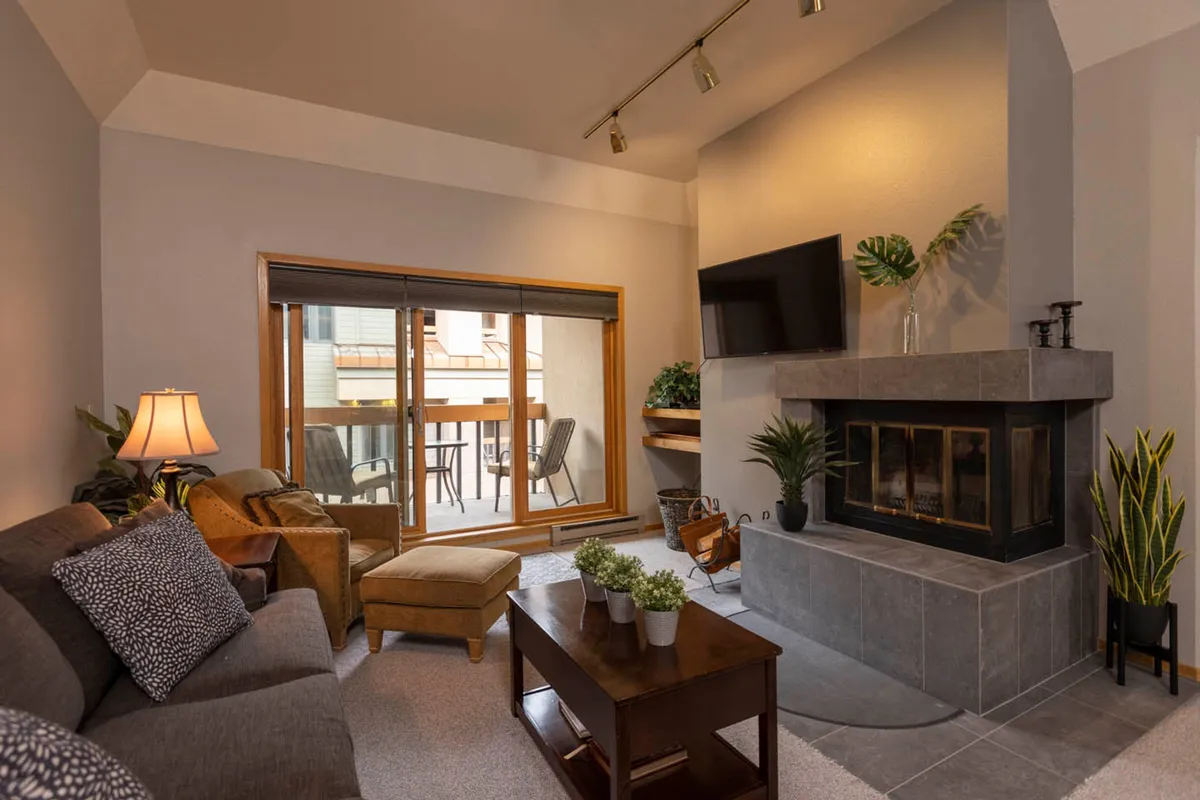 The image showcases a warm, inviting living room with a crackling fireplace, plush sofa, and large windows opening onto a private balcony. Lush plants add a touch of nature, while the TV and cozy seating create an ideal setting for relaxing after a day on the slopes. The balcony promises mountain views, perfect for morning coffee or evening unwinding.