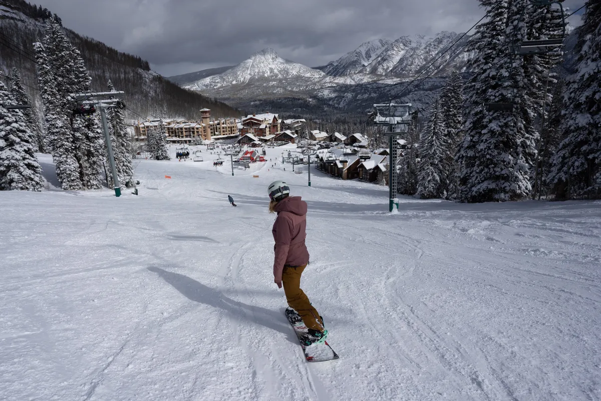A dramatic exterior perspective of a snowboarder descending powdery mountain slopes, with the ski resort, snow-dusted pine trees, and ski lifts visible against a backdrop of towering mountains.