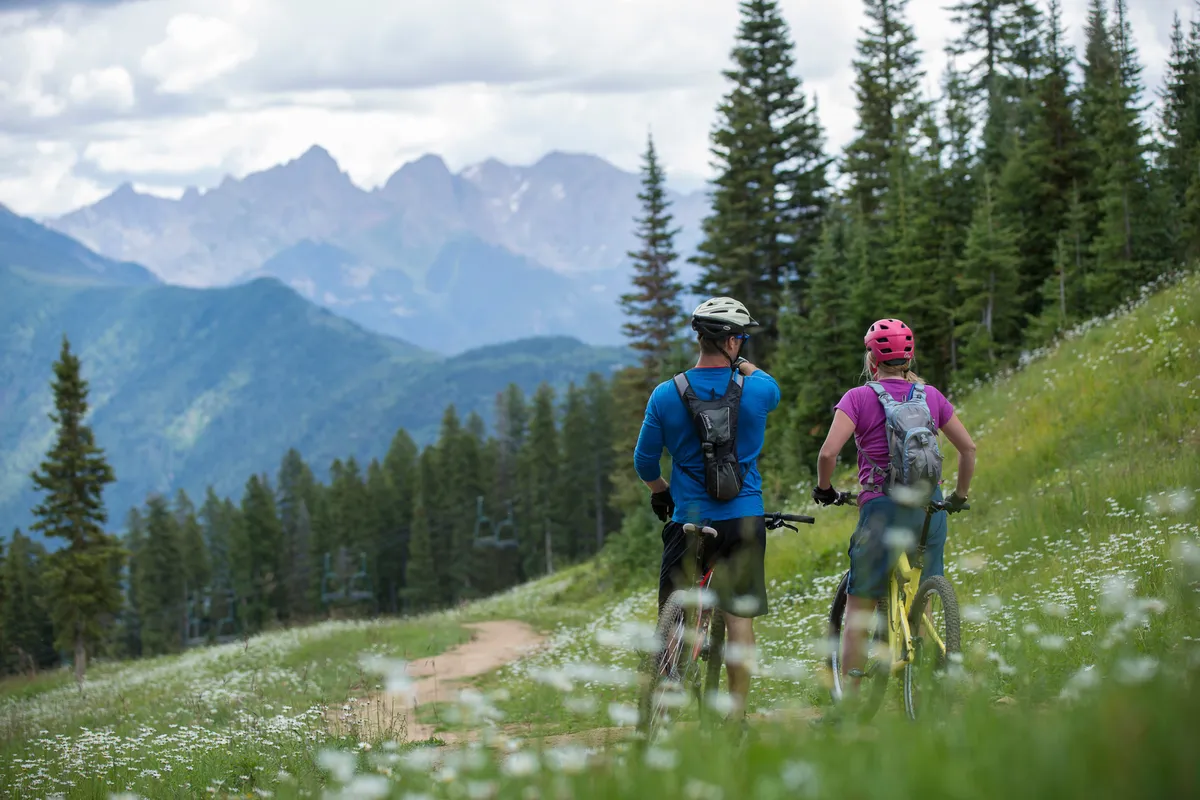 This exterior image features a scenic dirt trail winding through fields of wildflowers, with bicyclists riding against a breathtaking mountain range backdrop. The vibrant colors and active scene evoke the adventurous spirit of summer and off-season mountain escapes.