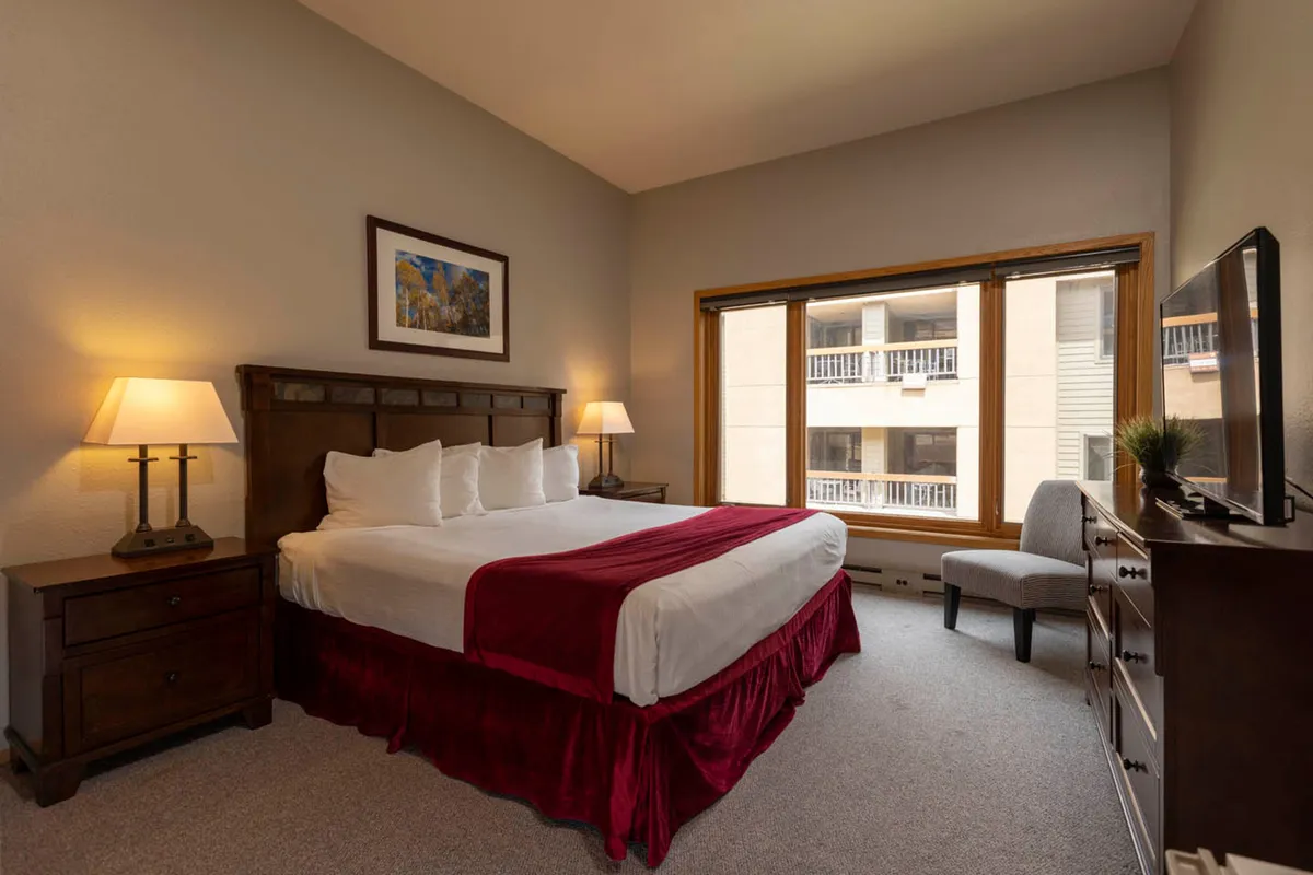 A comfortable bedroom with a bed adorned with a red accent, large windows providing views and light, a nightstand with a lamp, and a TV setup. The room exudes a restful, inviting ambiance, ideal for lazy mornings or evenings of relaxation.